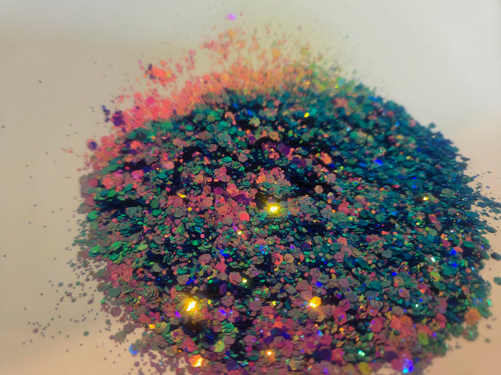 A pile of iridescent holographic glitter flakes with a rainbow of colors reflecting blue, green, pink, purple, and gold on a plain light background.