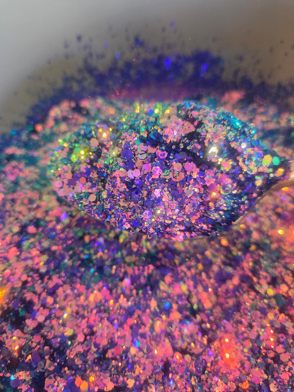 Close-up of iridescent holographic glitter in a mix of purple, pink, blue, green, and orange, with a small pile in the center and loose glitter scattered around on a light background.