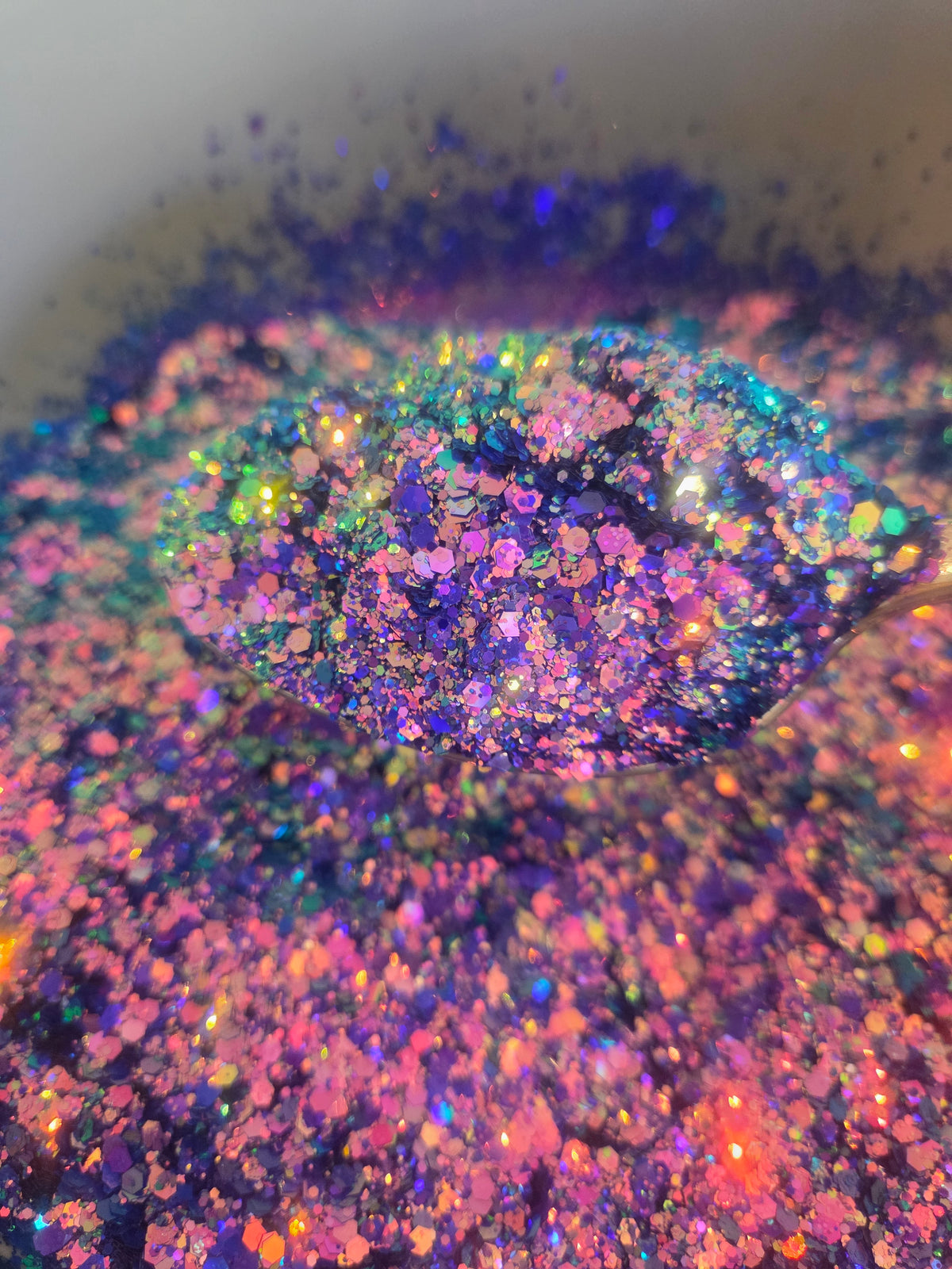 Close-up of iridescent holographic glitter in a mix of purple, pink, blue, green, and orange, with a small pile in the center and loose glitter scattered around on a light background.